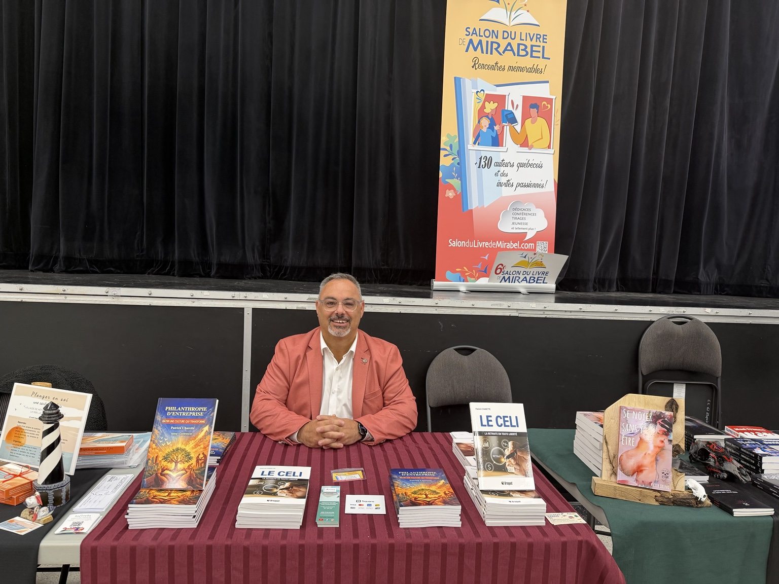 Patrick Charette au Salon du livre de Mirabel, entouré de ses ouvrages. Patrick Charette assis derrière son kiosque au Salon du livre de Mirabel, avec ses livres et la bannière officielle en arrière-plan.